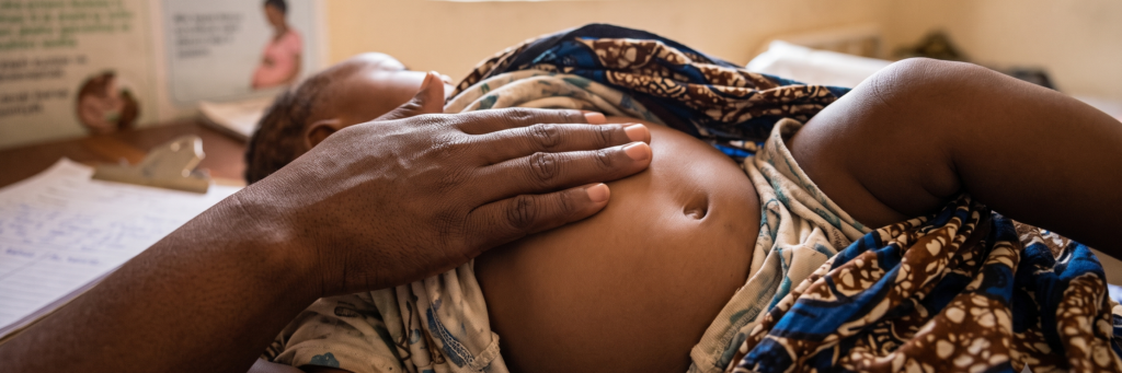 A close-up shot of an infant lying on a table in a health clinic setting. The baby's shirt is lifted to reveal their bare belly, where a mother's hand gently rests. Medical posters and clinic paperwork are visible in the background.