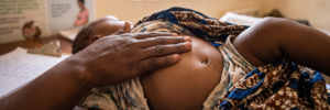 A close-up shot of an infant lying on a table in a health clinic setting. The baby's shirt is lifted to reveal their bare belly, where a mother's hand gently rests. Medical posters and clinic paperwork are visible in the background.