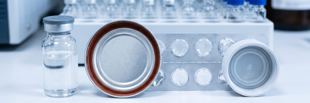 A clean close-up of everyday packaging under lab conditions: a plastic bottle cap, a blister pack, a food can lining and a glass vial with a rubber septum, arranged on a white bench next to an autosampler tray. Shallow depth of field, cool light.